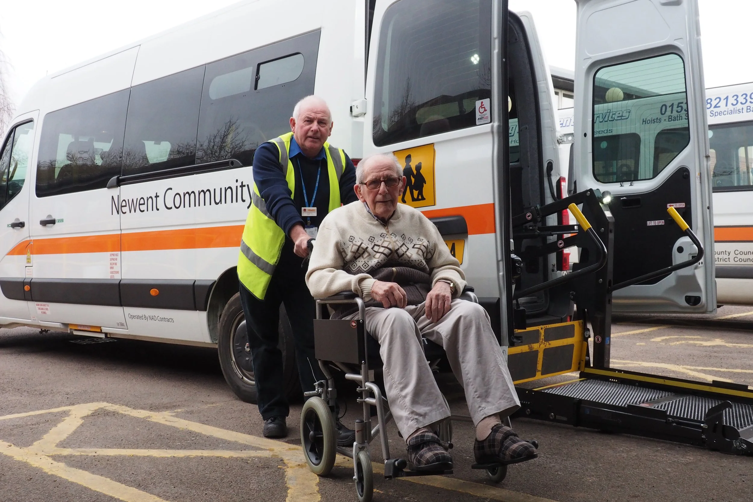 An elderly man sitting in a wheelchair being assisted by a community transport worker as he departs a minibus labeled 'Newent Community.' The worker is wearing a high-visibility vest and standing beside the man near the accessible lift of the bus.