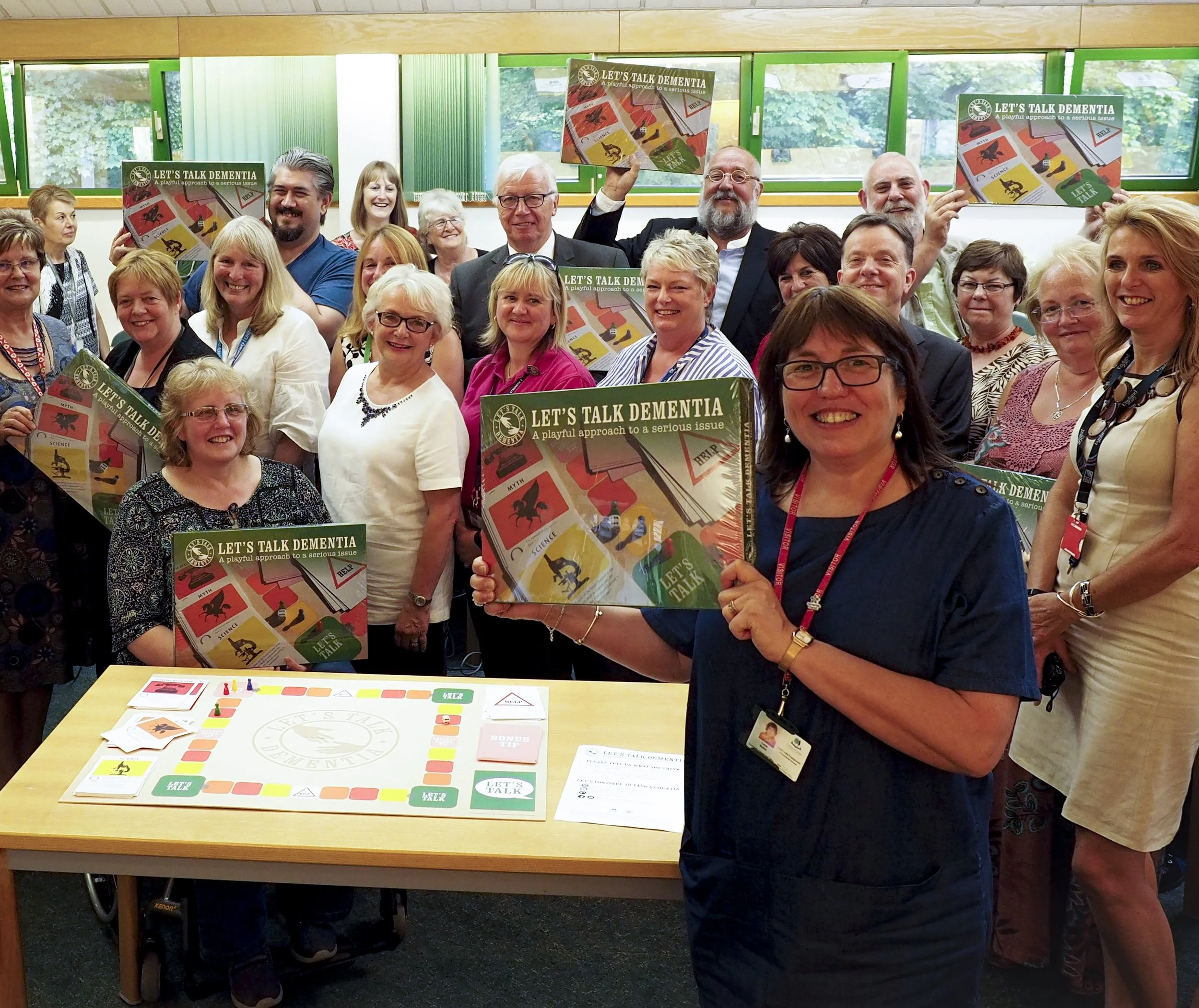 A group of people gathered in a room, some holding up board game boxes titled 'Let's Talk Dementia'. The game features colorful illustrations and text related to dementia awareness. The participants are smiling and appear to be at an event focused on dementia education or awareness.