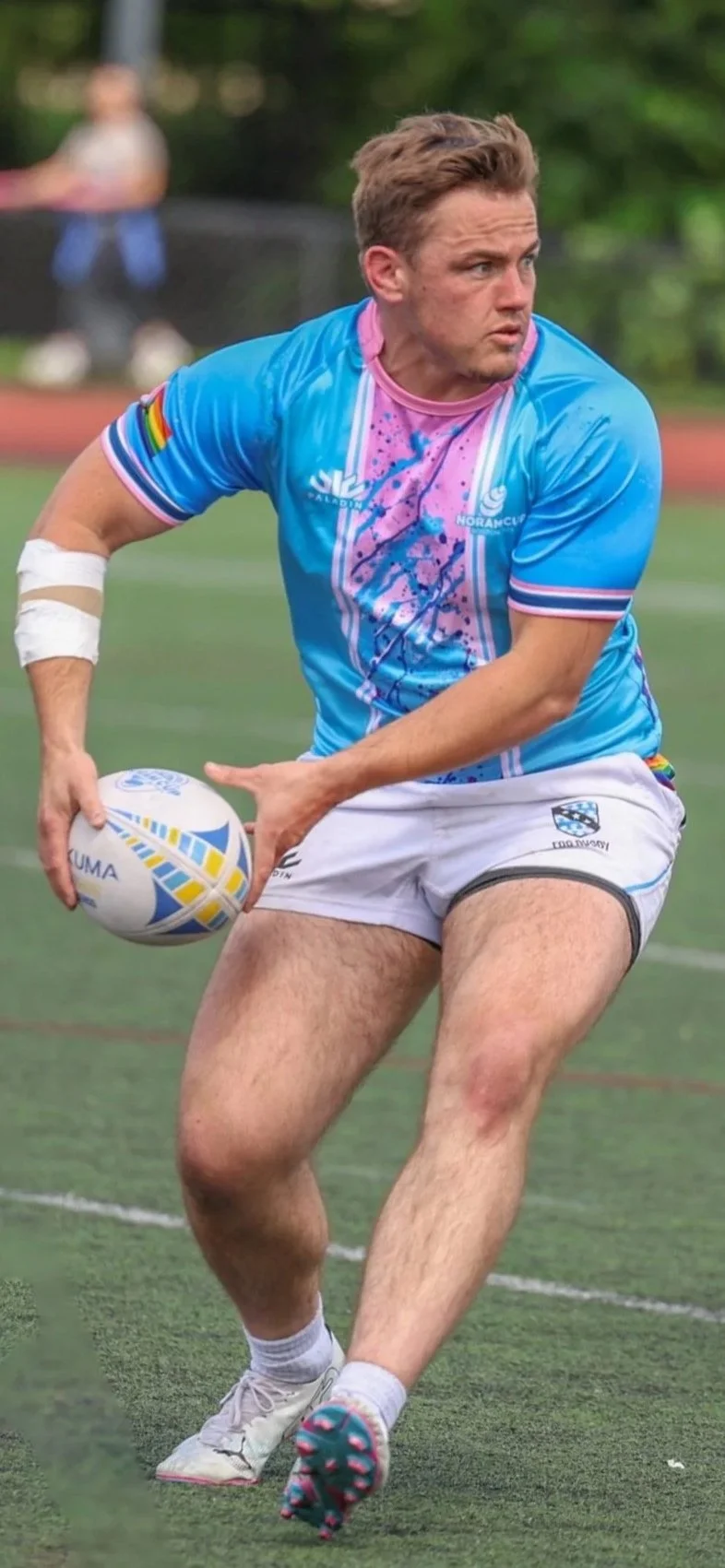 A rugby player in a blue and pink jersey holding a rugby ball on a field.