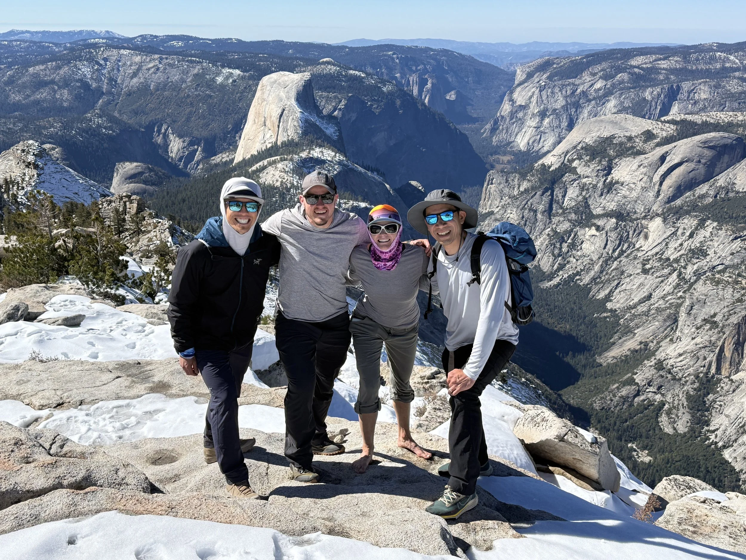 Four people standing on a snowy rock ledge, smiling, with a mountain landscape and Half Dome in the background.