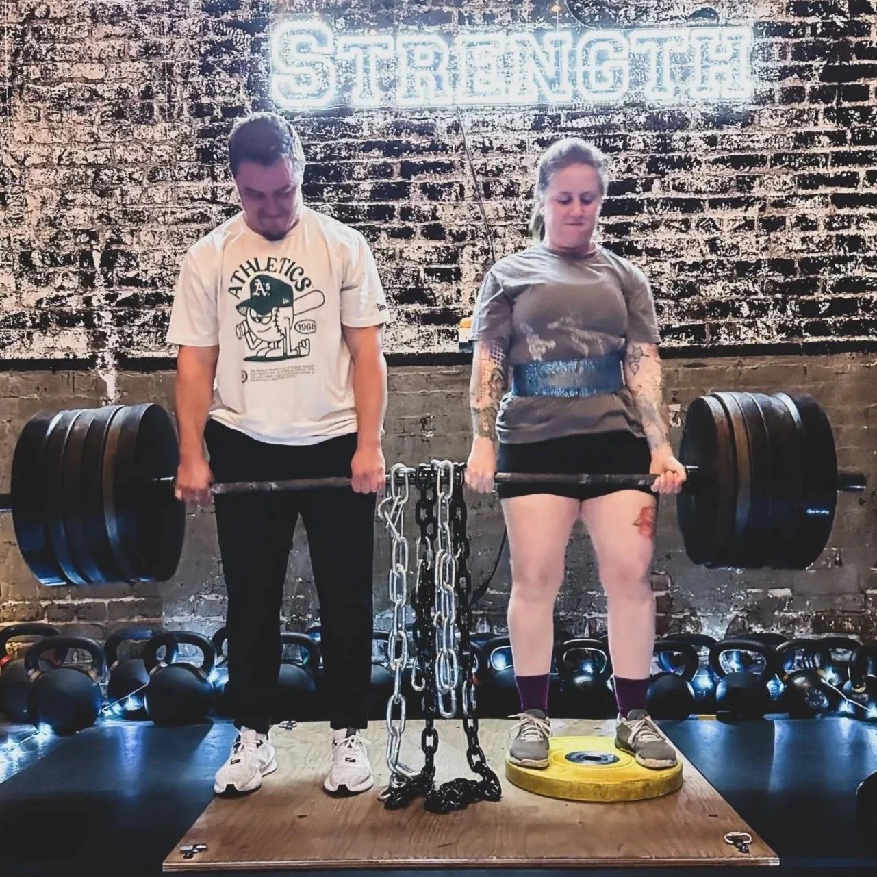 Two women standing on a wooden platform in a gym, lifting heavily loaded barbells. The woman on the left wears a white t-shirt and black pants, and the woman on the right wears a gray t-shirt, shorts, and sneakers. Behind them, there are kettlebells lined up on the floor and a chain hanging from their sides, with a neon sign above displaying the word 'STRENGTH' against a brick wall.