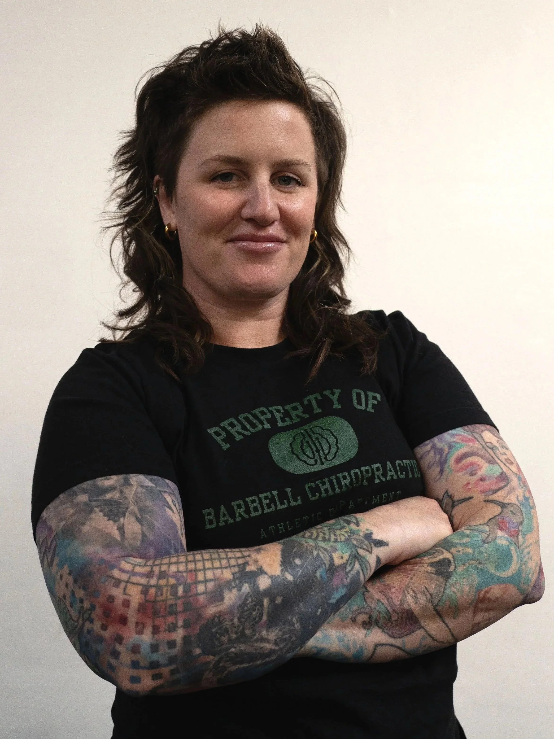 A woman with short, dark, wavy hair standing with arms crossed, wearing a black t-shirt with green text and colorful tattoos on both arms.