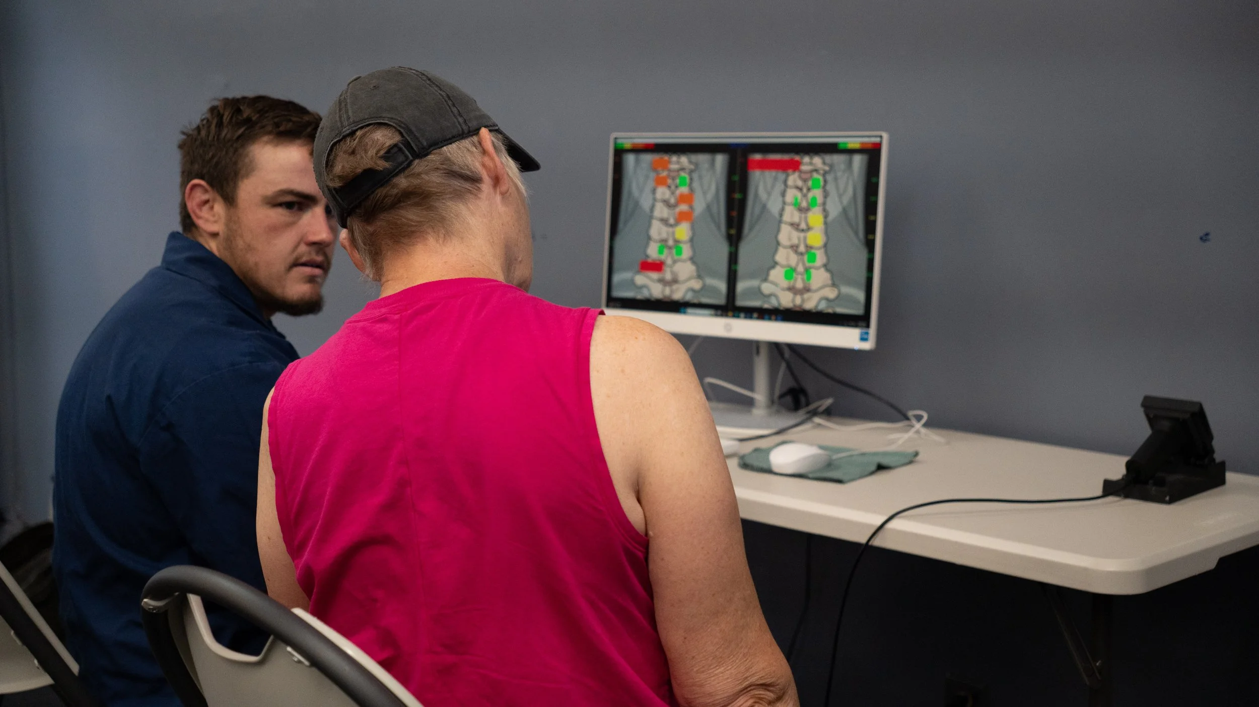 A woman in a pink sleeveless shirt sits in front of a computer screen displaying chiropractic images of a spine with color-coded markers, with a male chiropractor in a blue shirt beside her looking attentively.