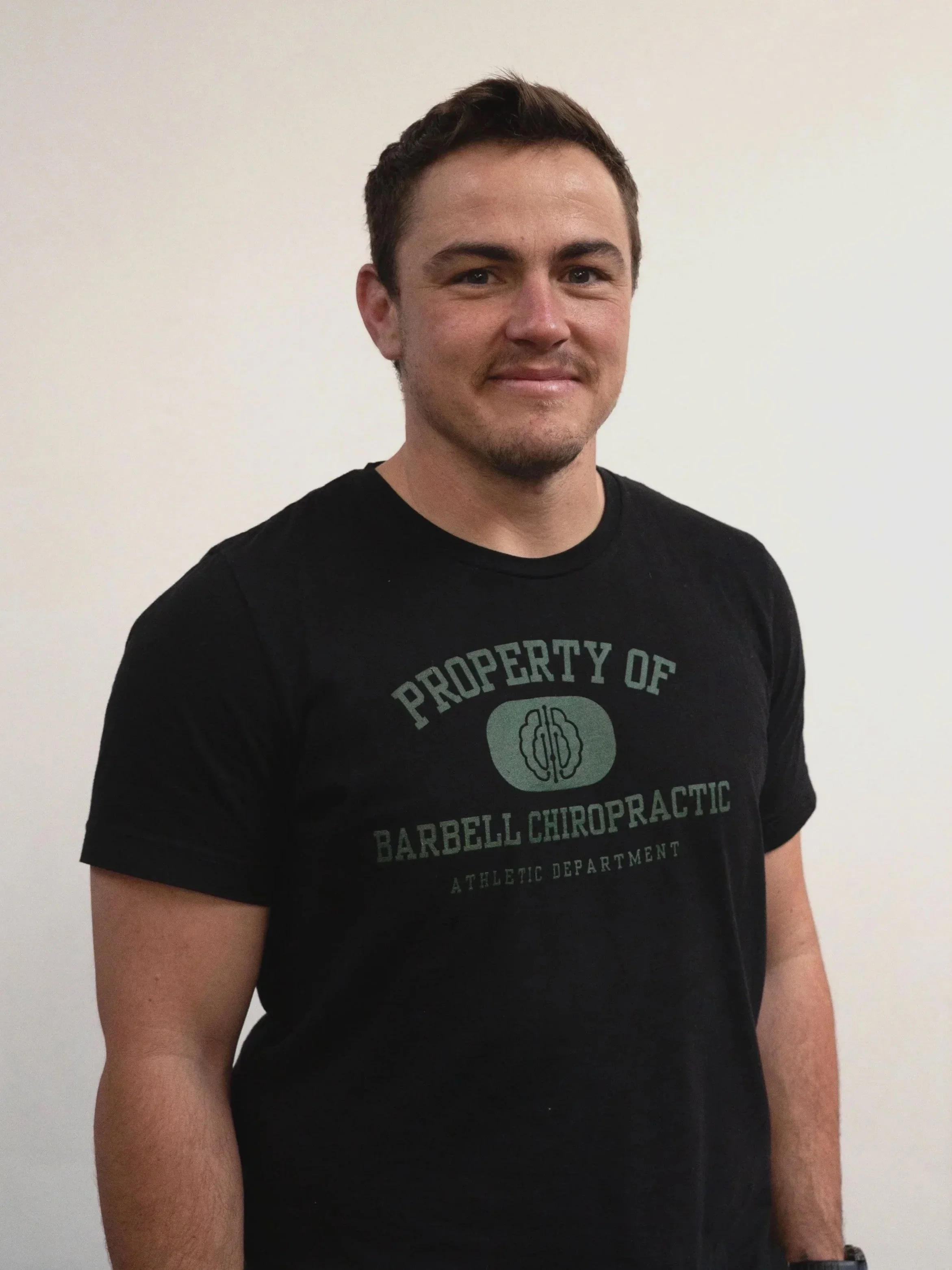 A man smiling wearing a black T-shirt that says 'Property of Barbell Chiropractic Athletic Department'.