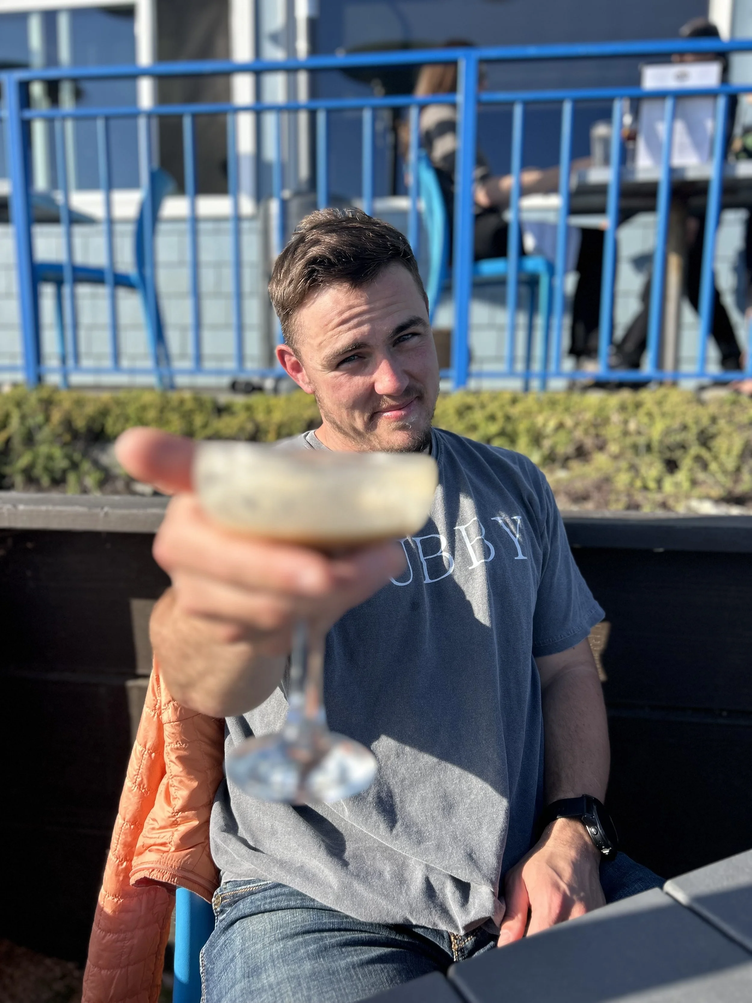 A man in a gray t-shirt and jeans extending a cocktail glass towards the camera, sitting outdoors at a table.