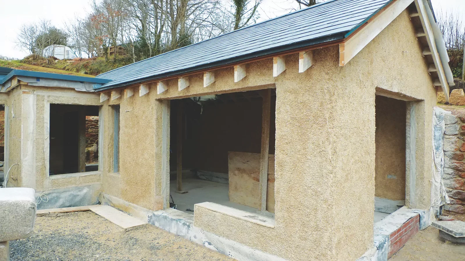 Construction site of a small building with unfinished walls and a metal roof, surrounded by dirt and construction materials.