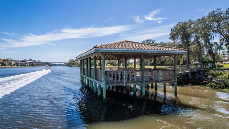 A wooden pavilion with a tiled roof extends over a waterway, with trees and residential buildings in the background and 