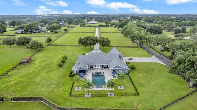 Aerial view of a large suburban home with a swimming pool, surrounded by a fenced yard with palm trees and open green fi