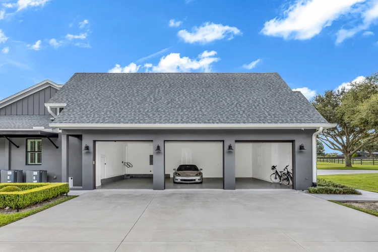 A modern three-car garage with one car and two bicycles parked inside, gray exterior, with a concrete driveway, neatly t
