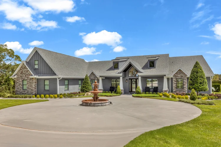 Large modern house with gray siding, brick accents, and a gabled roof, featuring a circular driveway, a fountain, and la