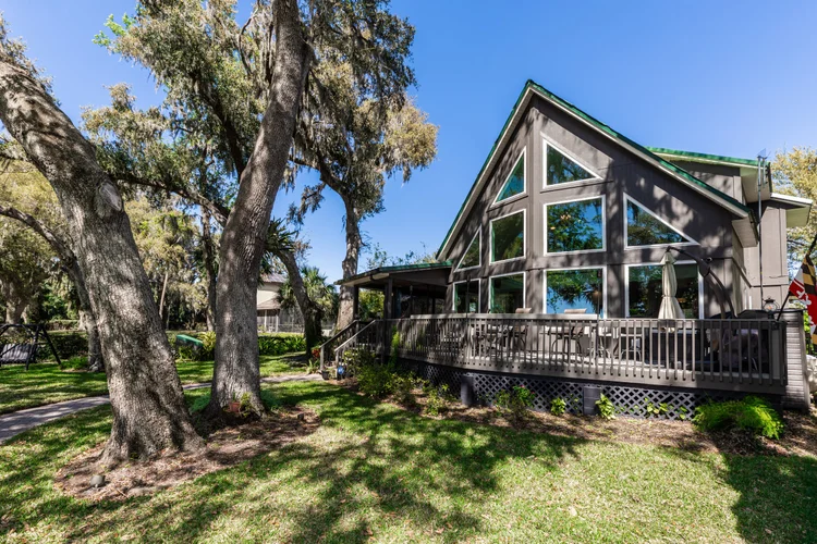 A house with large triangular windows, a wooden deck, and a lush green yard with tall trees and grass on a sunny day.