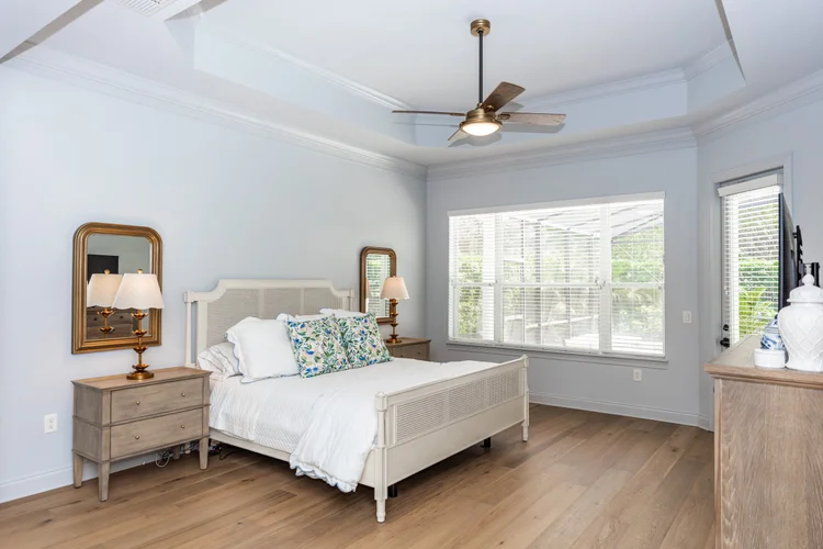 A bedroom with a white bed, wooden nightstands with lamps, a dresser with decorative items, a ceiling fan, large window 
