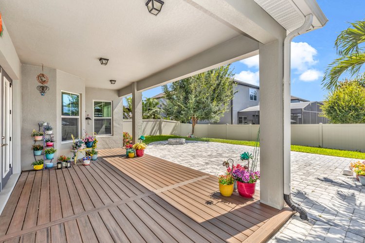 Backyard patio with wooden deck, potted colorful flowers, a tree, a fire pit, and a stone-paved area, enclosed by a ligh