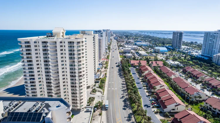Aerial view of a coastal city with tall white apartment buildings, a road, and beachfront houses by the ocean on a clear