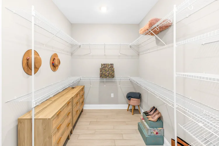 Empty walk-in closet with white wire shelving, a wooden dresser, pink high heels, hats on the wall, and boxes on the flo
