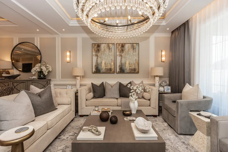Elegant living room with a central beige sofa, gray and dark cushions, a large chandelier overhead, and a coffee table w