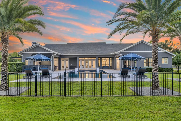 A modern house with a backyard featuring a swimming pool, poolside chairs with umbrellas, palm trees, and a sunset sky.