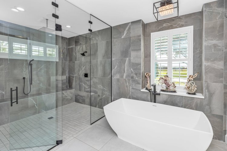 Modern bathroom with large gray tiled walk-in shower, white bathtub, window with decorative figurines, and contemporary 