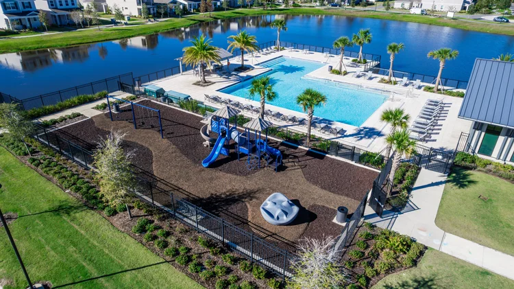 An aerial view of a community pool area with palm trees, lounge chairs, and a playground, situated next to a lake with r