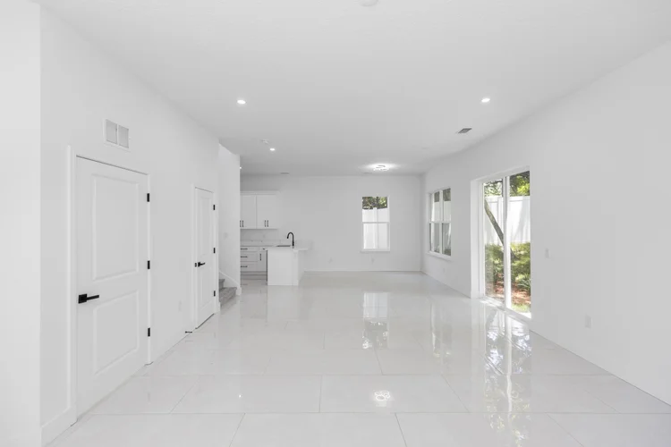 Empty white living room with large tiled floor, multiple windows, glass sliding door to outside, and a corner kitchen wi
