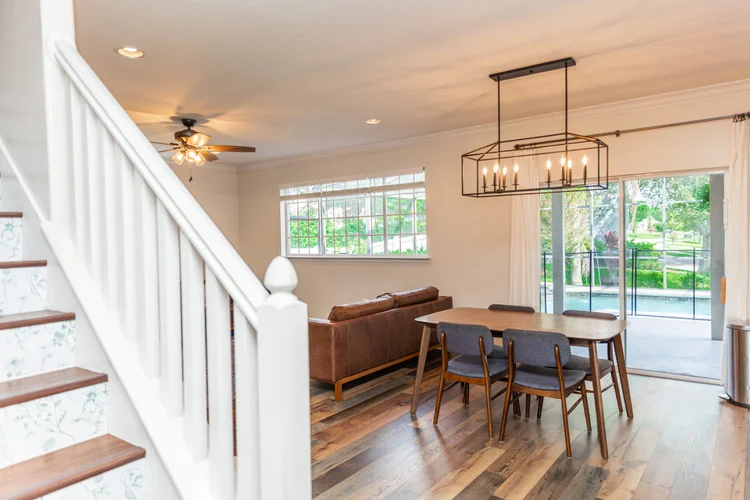 Living room with wooden dining table and chairs, brown leather sofa, sliding glass door, windows, ceiling fan, chandelie