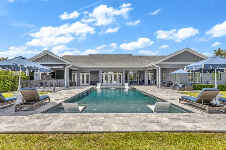 A backyard with an in-ground swimming pool surrounded by lounge chairs with blue cushions, umbrellas, and a patio with o