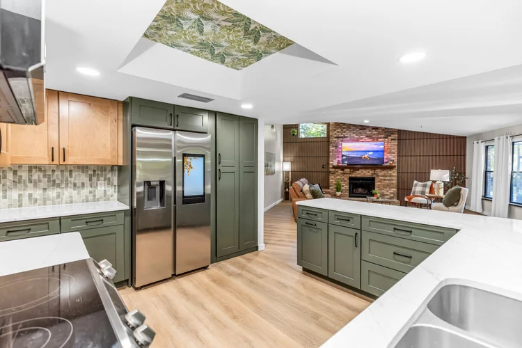 Modern kitchen with green cabinets, stainless steel refrigerator, and white countertops, opening to a living room with b