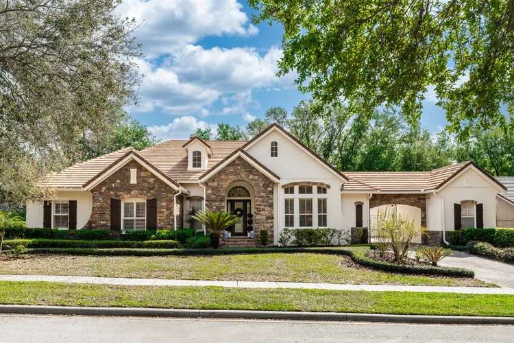 Front view of a large suburban house with stone and white exterior walls, a tiled roof, and well-maintained lawn and tre