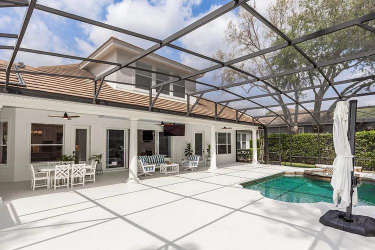 Backyard patio with pool, enclosed by a screened structure, with outdoor furniture, potted plants, and a house with whit