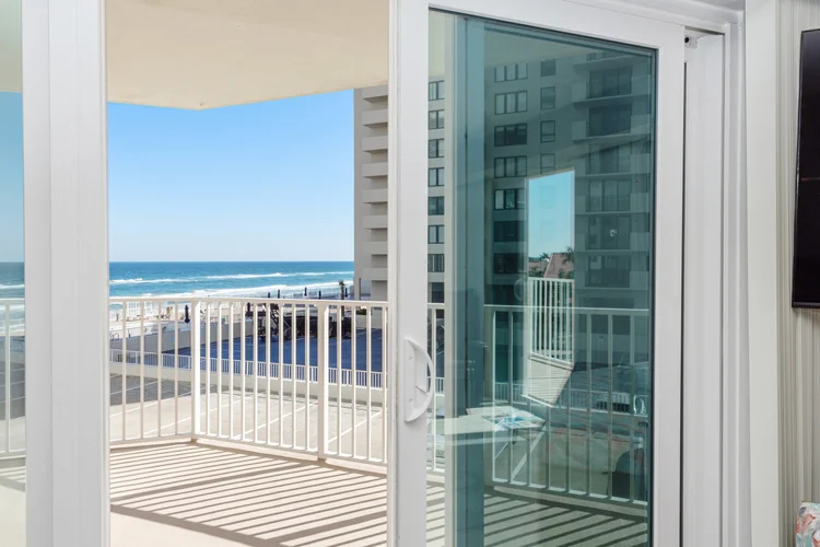 Balcony with white railing overlooking the beach and ocean, with a neighboring building in the background, seen through 