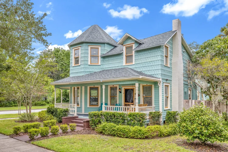 A two-story, Victorian-style house painted light blue with decorative shingles, a wraparound porch, and a turret, surrou