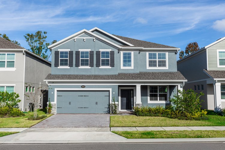 A two-story blue house with black shutters, a gray garage door, and a small porch, surrounded by green grass and bushes,
