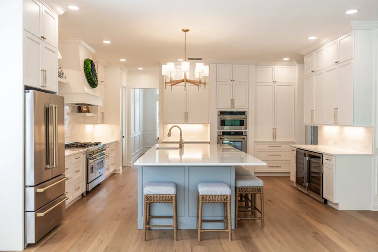 Modern white kitchen with a large island, stainless steel appliances, and wooden floors. Central chandelier hanging abov
