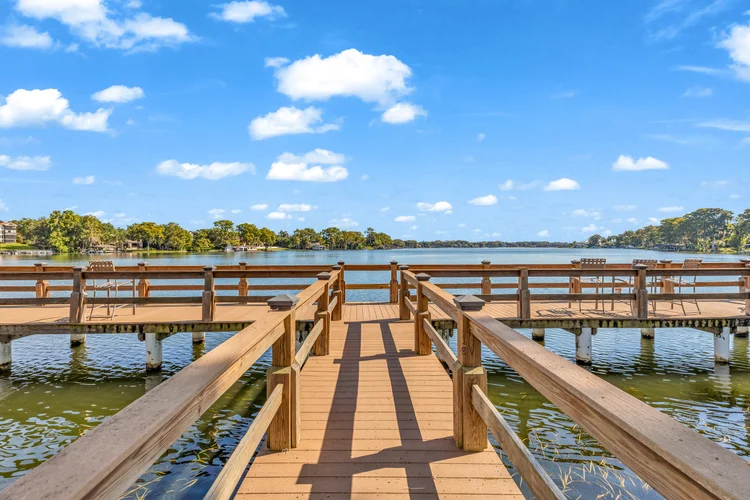 A wooden dock extending over a lake, with chairs on the sides, under a blue sky with scattered white clouds.