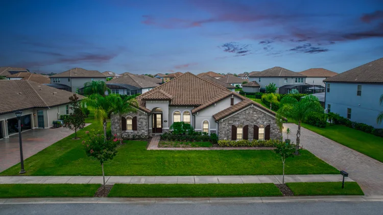A large modern suburban house with a well-manicured lawn, stone and stucco exterior, and tan tile roof, situated in a ne
