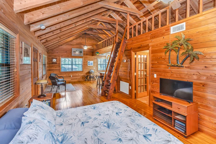 Interior view of a wooden cabin living room with a sloped ceiling, a bed in the foreground, a TV on a wooden stand, a se