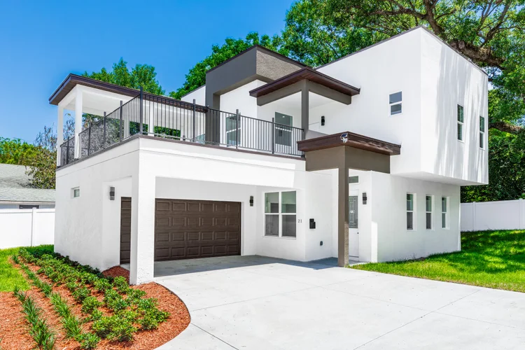 Modern white two-story house with a brown garage door, a small front yard with green grass and plants, a balcony with bl