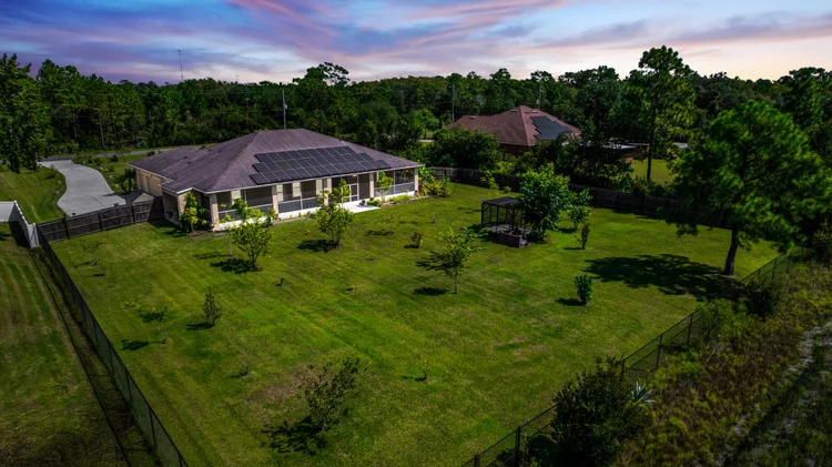 A house with solar panels on the roof, surrounded by a large fenced backyard with several small trees, a porch, and a ga