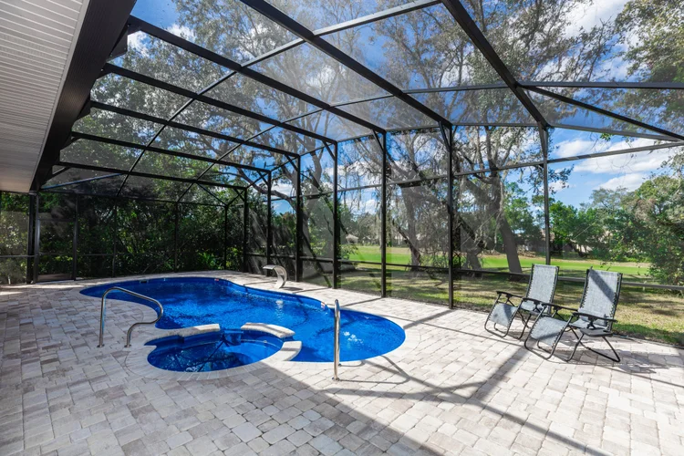 Enclosed backyard swimming pool area with two lounge chairs, a hot tub, and a screen enclosure with a view of trees and 