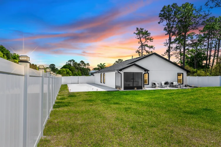 Backyard with a white fence, a concrete patio, a white house with black trim, outdoor seating, and tall trees at sunset.