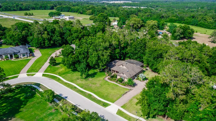 An aerial view of a suburban neighborhood with houses, green lawns, and trees, with a highway in the background.