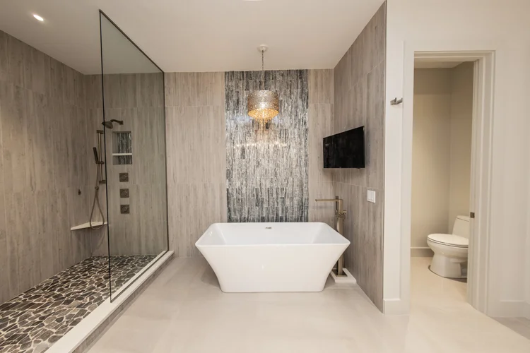 Modern bathroom with walk-in shower with pebble tile floor and grey stone walls, white bathtub with sleek brass faucet, 