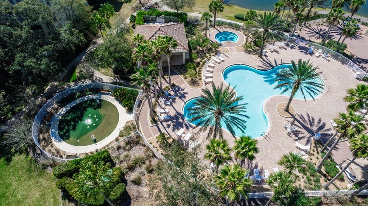 Aerial view of a swimming pool area with multiple pools, surrounded by palm trees, lounge chairs, and a small building, 