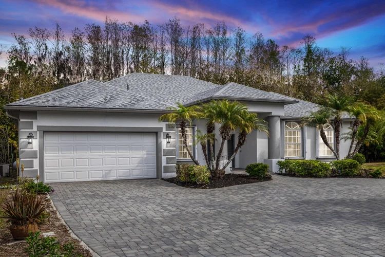 White single-story house with a gray shingle roof, two garage doors, palm trees near the entrance, landscaped bushes, an