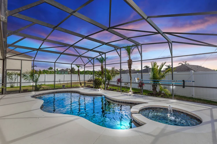 A backyard swimming pool enclosed by a screen structure during twilight, with a hot tub and tropical plants, a white fen