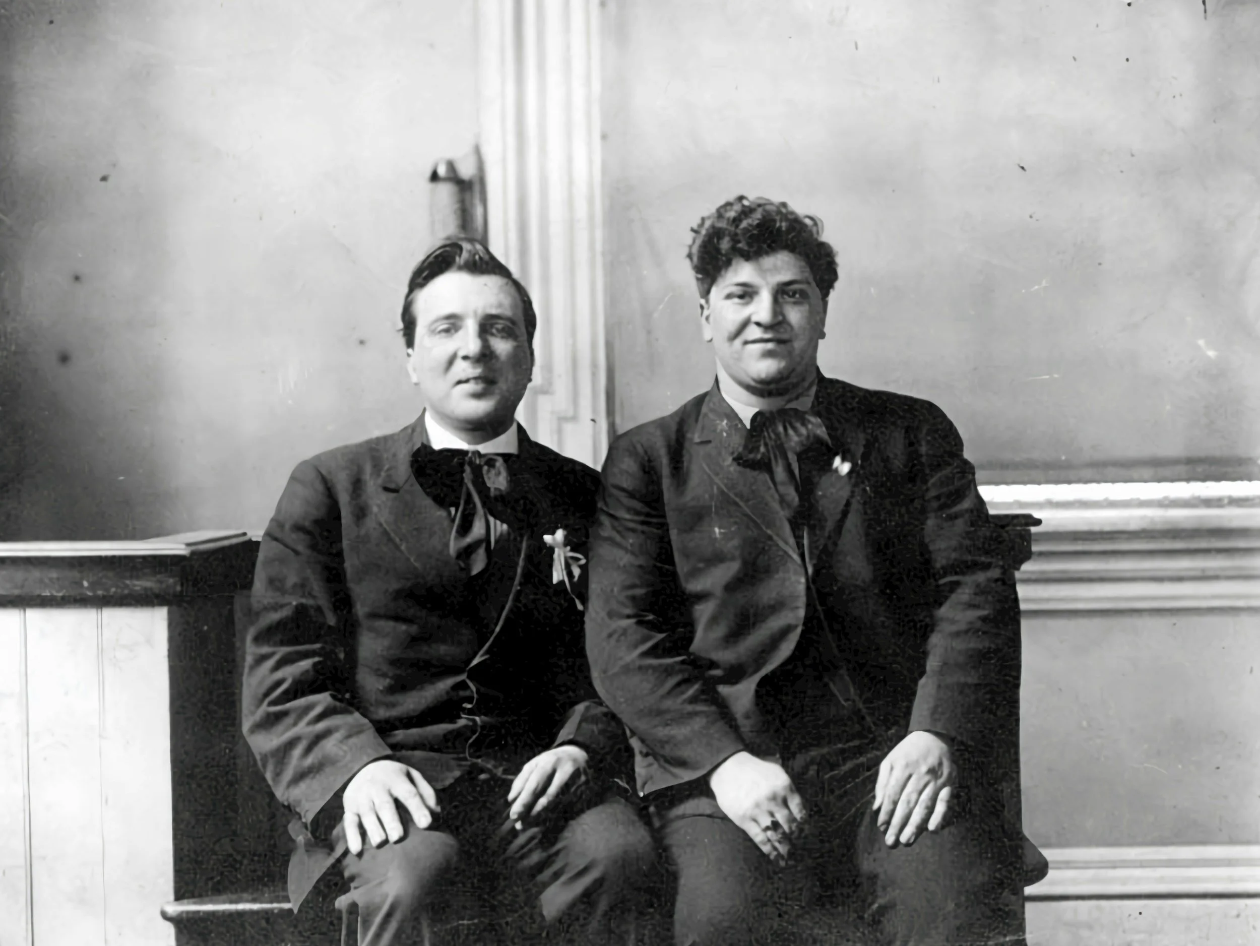 A black and white photo of two queer people dressed in formal wear sit next to each other.