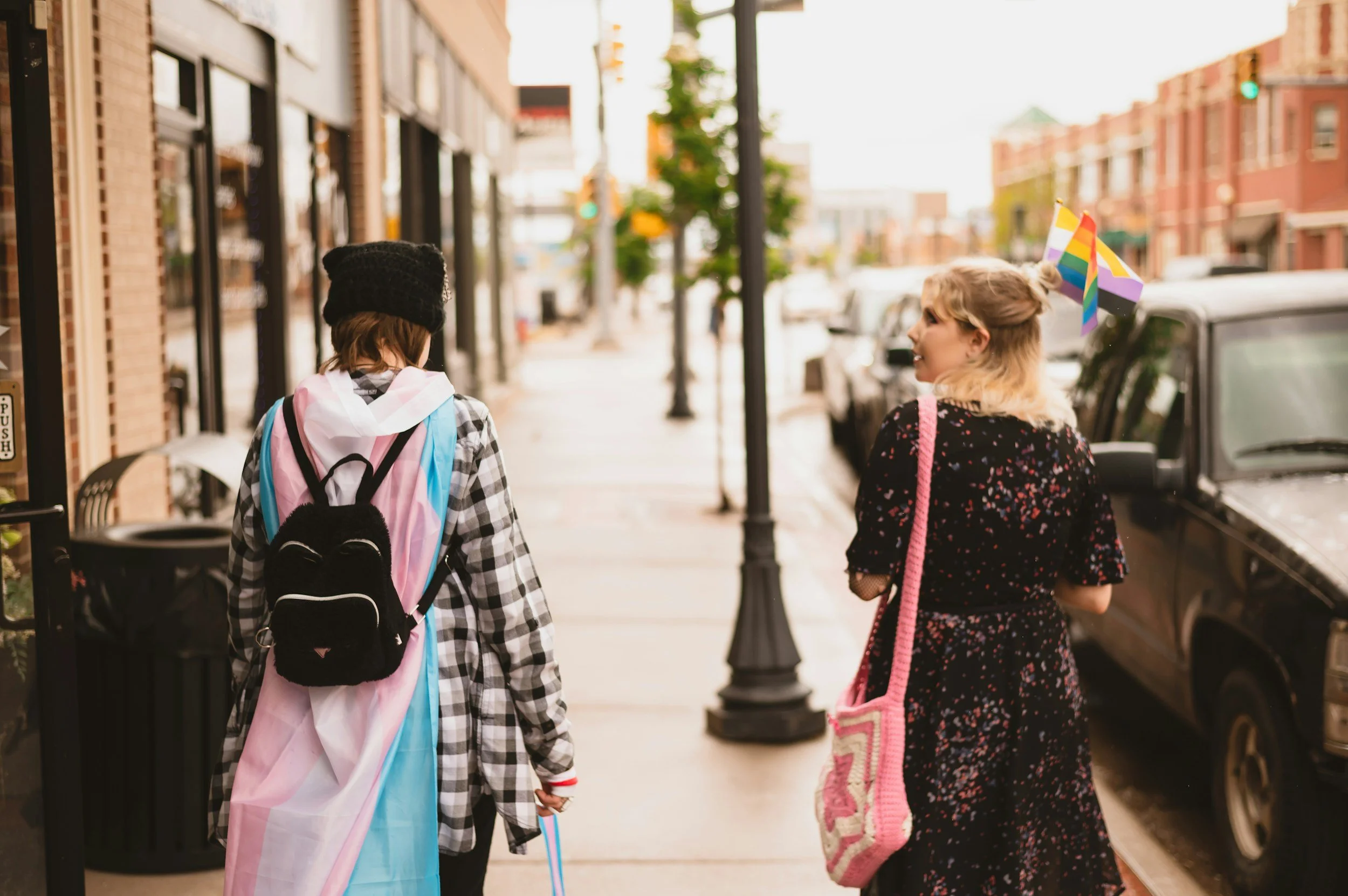 Two teenagers walking outside on a city street. One wears a transgender flag as a cape. The other has a lesbian flag and a pride flag in their hair.
