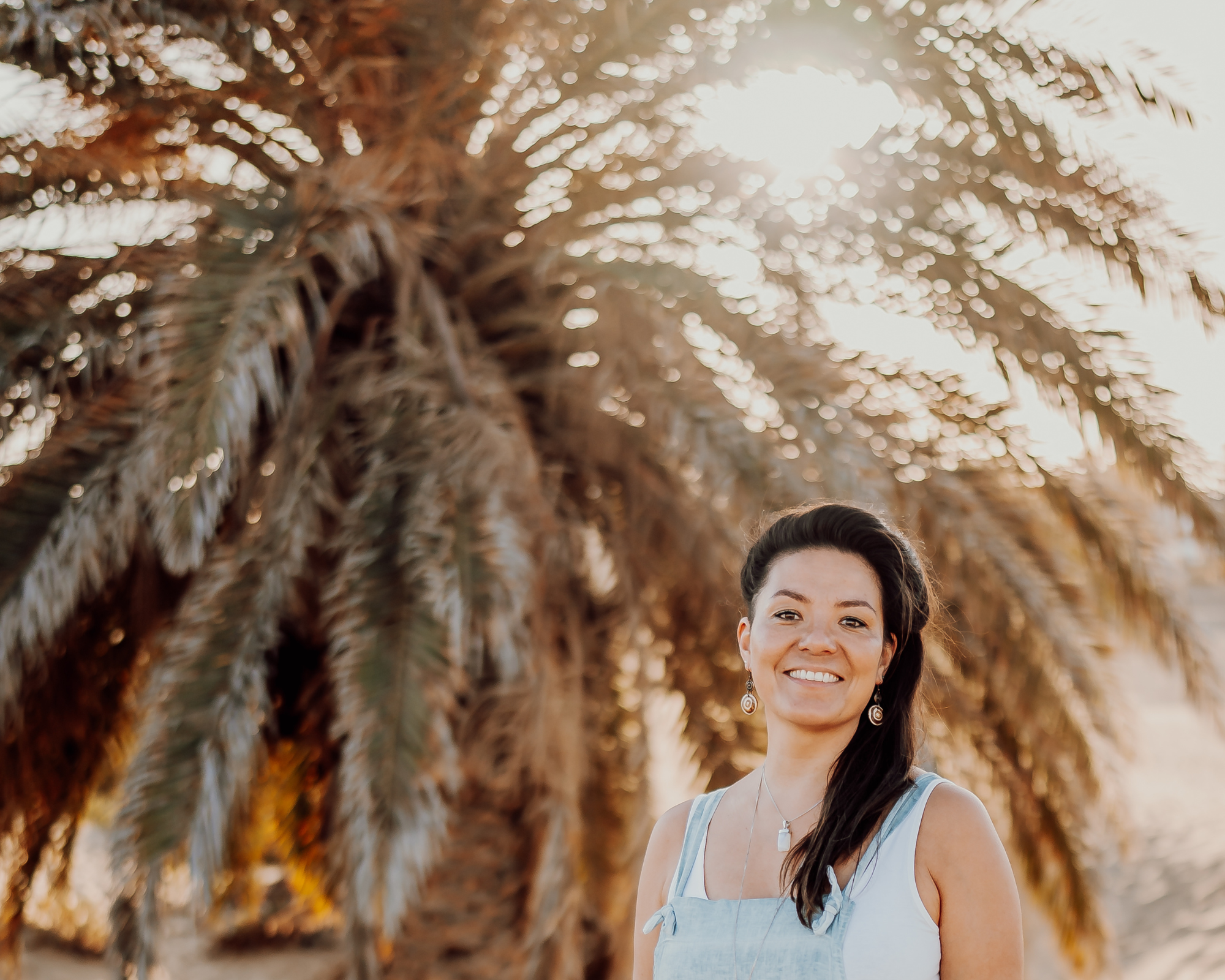 A woman smiling outdoors with palm trees in the background, sunlight shining through the trees.