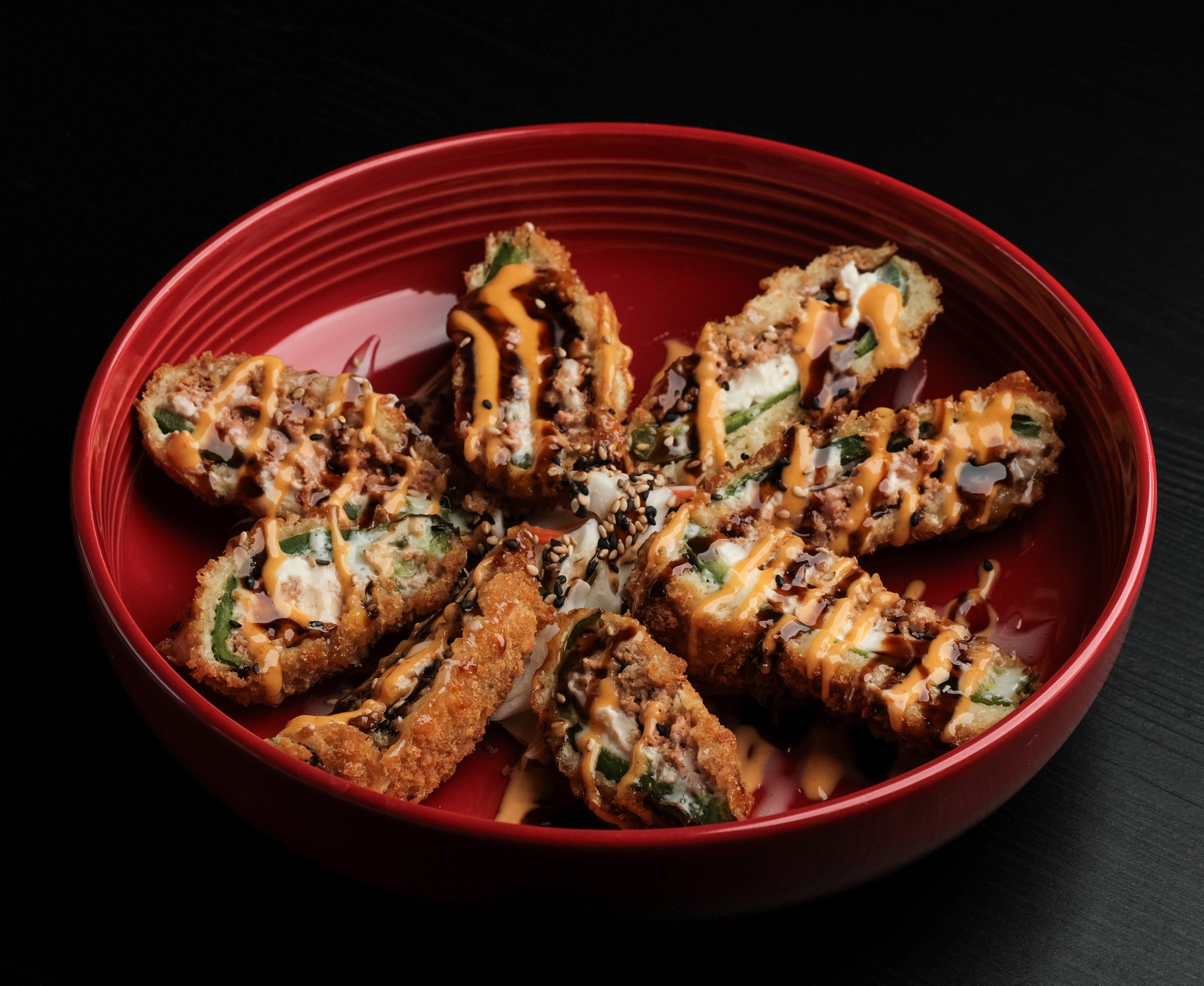 A red bowl with fried sushi rolls topped with drizzle of spicy mayo and eel sauce, garnished with sesame seeds, on a black surface.