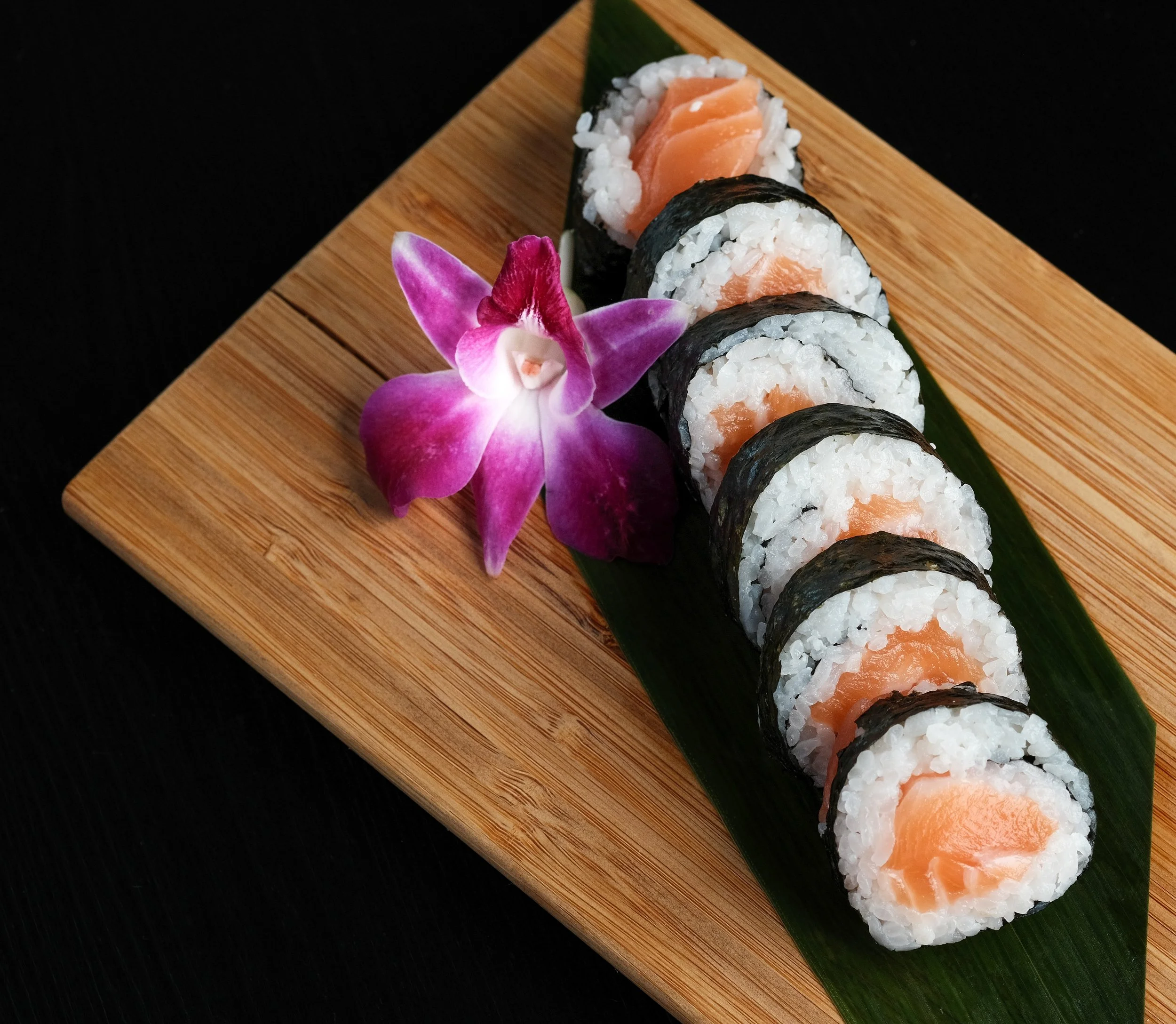 A wooden platter with five pieces of salmon sushi rolls arranged in a line on a green leaf, decorated with a pink and purple orchid flower, against a black background.